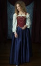 A Renaissance woman wearing a red brocade corset and blue silk skirt and standing before an ornately decorated backdrop