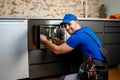 Young repairman in a blue uniform repairs and repairs a microwave oven with a screwdriver in a modern kitchen Royalty Free Stock Photo