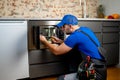 Young repairman in a blue uniform repairs and repairs a microwave oven with a screwdriver in a modern kitchen Royalty Free Stock Photo