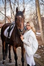 Young rider woman blonde with long hair in white clothes standing near horse Royalty Free Stock Photo