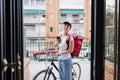 Young rider woman wearing red backpack delivering food on a bike, checking order with smart phone while standing on street gate in Royalty Free Stock Photo