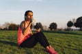 Young runner woman seated in the grass before run in a park, drinking water with smartphone and earphones. Close up athletic and