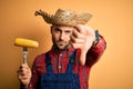 Young rural farmer man holding countryside corn over isolated yellow background with angry face, negative sign showing dislike