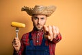 Young rural farmer man holding countryside corn over isolated yellow background pointing with finger to the camera and to you,