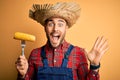 Young rural farmer man holding countryside corn over isolated yellow background very happy and excited, winner expression