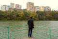 Young Russian guy in black coat with dark bag standing with his back to the camera near the lake thinking and looking at houses Royalty Free Stock Photo