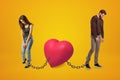 Young sad man and woman wearing casual clothes chained to pink heart on yellow background