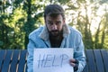 Young sad sick poor homeless man sitting on the bench on the street holding help sign paper feeling lonely and depressed because Royalty Free Stock Photo