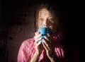 Young sad woman portrait behind the window in the rain with rain drops on it. girl holding a cup of hot drink Royalty Free Stock Photo