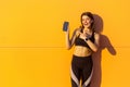 Young satisfied beautiful sporty woman in black sportwear standing near orange wall background and holding phone, pointing finger Royalty Free Stock Photo