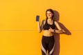 Young satisfied beautiful sporty woman in black sportwear standing near orange wall background and holding phone, showing thumbs Royalty Free Stock Photo