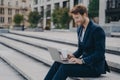 Young satisfied businessman in stylish suit sitting on stairs outside with laptop and coffee to go Royalty Free Stock Photo