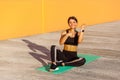 Young satisfied sporty girl in black sporwear in morning on street, sitting on green mat, pointing finger and showing good result Royalty Free Stock Photo