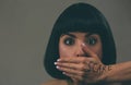 Young scared woman with black hair posing on camera. Scared afraid model look straight. Mouth closed with hand. Fear in