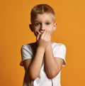 Young school boy kid in white t-shirt stands scared, eyes wide opened in surprise, covering his mouth with both hands