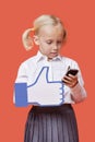 Young schoolgirl with cell phone holding fake like button against orange background