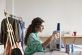 Young seamstress with dark curly hair in colorful shirt dreamily using sewing machine in sewing workshop