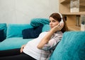Young serene charming pregnant woman enjoying classical music on headphones, relaxing on the couch at home during her maternity