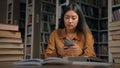 Young serious asian woman student sitting in library at desk searching information on Internet using mobile phone Royalty Free Stock Photo