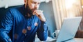 Young bearded businessman sitting in front of computer, looking at screen, holding pen, thinking. Freelancer works home.