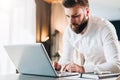 Young serious bearded businessman standing in office near table and using laptop. Man works on computer, checks e-mail Royalty Free Stock Photo