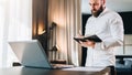 Young serious bearded businessman stands near desk opposite laptop and makes notes in notebook. Student is studying