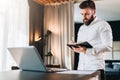 Young serious bearded businessman stands near desk opposite laptop and makes notes in notebook. Student is studying