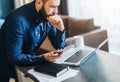 Young serious bearded businessman working on computer, holding smartphone, thinking. Man analyzes information, planning.