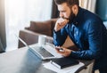 Young serious bearded businessman working on computer, holding smartphone, thinking. Man analyzes information, planning.