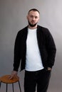 Young serious bearded man with short dark hair wearing black jacket, jeans, white T-shirt, standing at wooden stool. Royalty Free Stock Photo