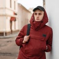 Young serious confident man in a red stylish long jacket with a hood in a black fashionable cap with a trendy sports backpack