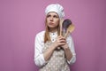 Young serious cook girl in apron holds spoons and kitchen items and looks at a place for text on a pink background, portrait of a