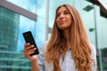 Young serious and determined business woman standing in front of modern building with phone in her hand