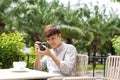 Young serious male sitting at wooden table with photo camera and preparing for photo session while having coffee break Royalty Free Stock Photo