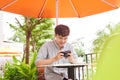 Young serious male sitting at wooden table with photo camera and preparing for photo session while having coffee break Royalty Free Stock Photo