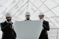 Young serious indian man in helmet and tie with tablet escorted with two engineers and pointing at building pipes. Controller Royalty Free Stock Photo