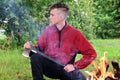 young serious man with a mohawk hairstyle is sitting with an axe by a fire in the forest. Take a break. Path to nature. Royalty Free Stock Photo