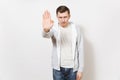 Young serious severe man in t-shirt, blue jeans and light sweatshirt showing stop gesture with palm, frowns on