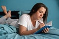 Young serious woman laying on bed and writing a diary. Caucasian lady with notebook and pen in her hands. Bright home Royalty Free Stock Photo