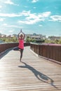 Young serious woman practicing yoga with hands raised up outdoors near on th wooden bridge against blue sky with clouds