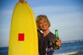 Young beautiful and happy surfer woman holding yellow surf board smiling cheerful drinking beer bottle enjoying summer holida