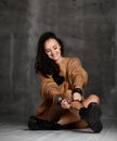 Young fashion woman sitting on floor in brown knitted blouse tying shoelaces on dark night Royalty Free Stock Photo