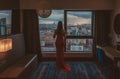 Young sexy girl in a long red dress on a background of the panoramic windows with views of the city from the hotel room