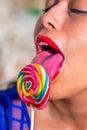 Young sexy Latina with bright red sensual lips passionately licking a colorful spiral lollipop with her beautiful sensual long Royalty Free Stock Photo