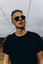 Young sexy man in black shirt and sunglasses standing outdoor. Dark key photo