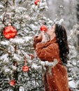 Young sexy brunette woman in stylish fur coat stands near decorated Christmas tree kisses red balls under snow