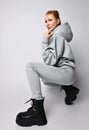 Young sexy slim red-haired woman in grey hoodie, pants and black massive shoes sits squatted side to camera