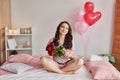 Young sexy woman on bed in pink pajamas with bouquet of red roses, room decorated with balloons. Postcard Happy Valentine`s Day, Royalty Free Stock Photo