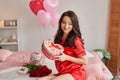 Young sexy woman on bed in red pajamas with heart-shaped gift box, room is decorated with balloons and red roses. Happy Valentine Royalty Free Stock Photo