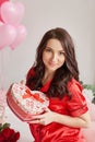 Young sexy woman on bed in red pajamas with heart-shaped gift box, room is decorated with balloons and red roses. Happy Valentine Royalty Free Stock Photo